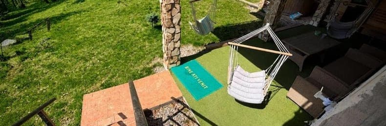 Terasa acoperită, cu un hamac-leagăn alb pe iarbă și ciubărul de lemn în capăt, deschisă spre pajiște și pădure.
